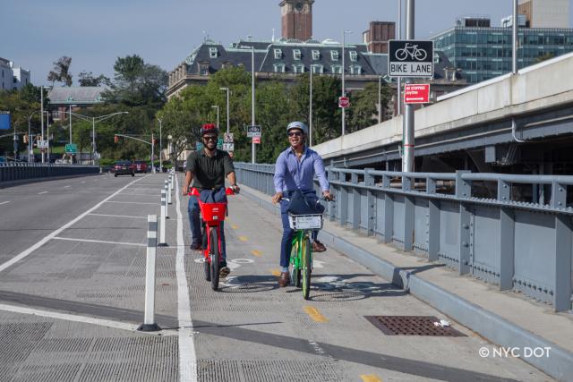 Two guys riding bikes, going towards the Staten Island Ferry Terminal. One man is in casual clothing and on a JUMP bike. The other man is in business-casual attire , riding a Lime Bike.