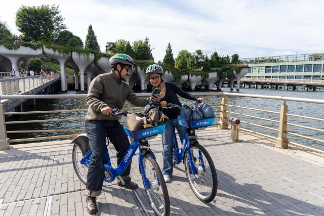 Man shows woman something on smart phone while both sitting on Citi Bikes.